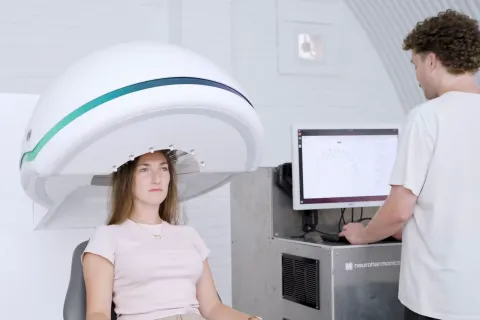 Woman sits under machine that wraps around her head while clinician stands next to her looking at computer