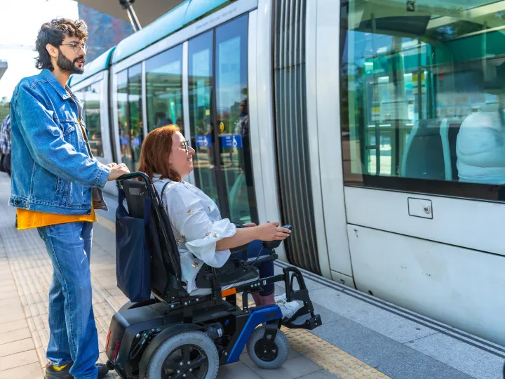 Man stands behind woman in wheelchair, as tram arrives