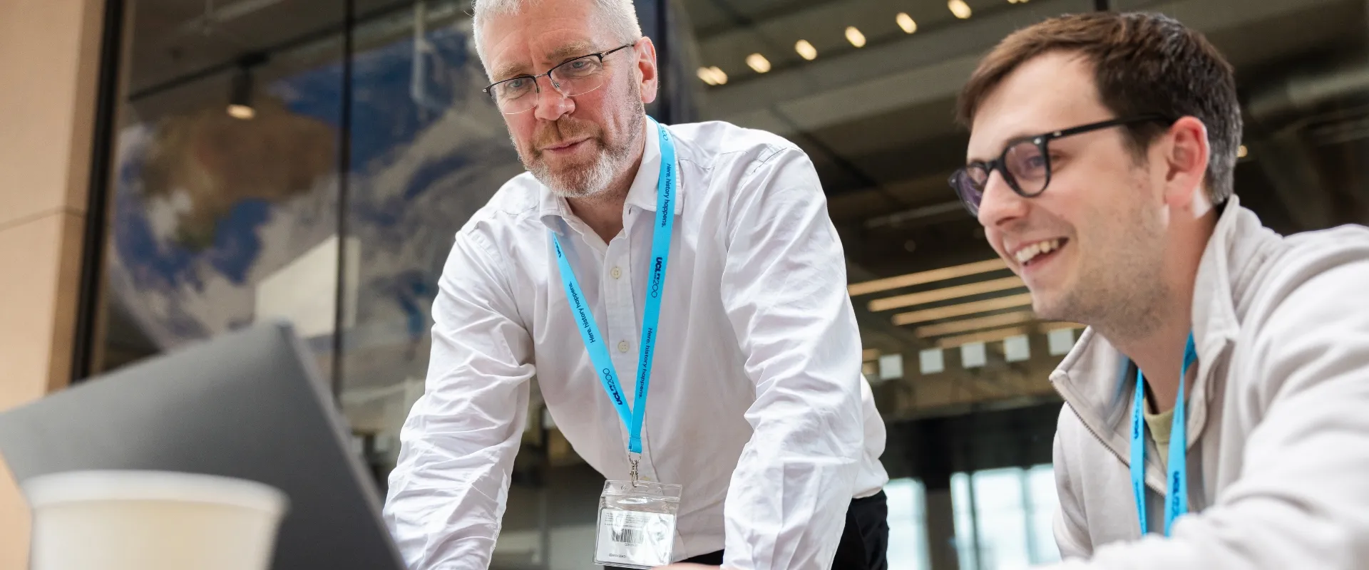 An older man in UCL lanyard leans forward onto the desk to look at a computer; younger man smiling working on the computer. 
