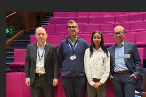 Four people stand in front of chairs in a lecture theatre. 