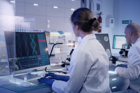 female scientist sits at computer looking at DNA; male colleague in background 