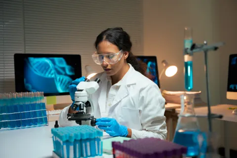 Woman researcher examining samples under microscope with digital DNA on screen, working on biotechnology and genetic discovery