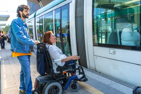 Man stands behind woman in wheelchair, as tram arrives