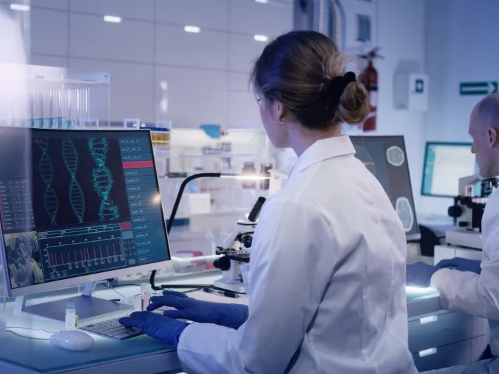 female scientist sits at computer looking at DNA; male colleague in background 