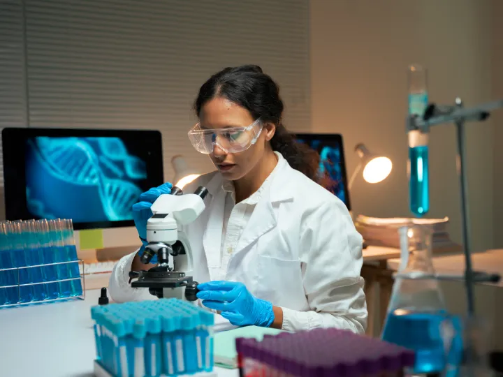 Woman researcher examining samples under microscope with digital DNA on screen, working on biotechnology and genetic discovery